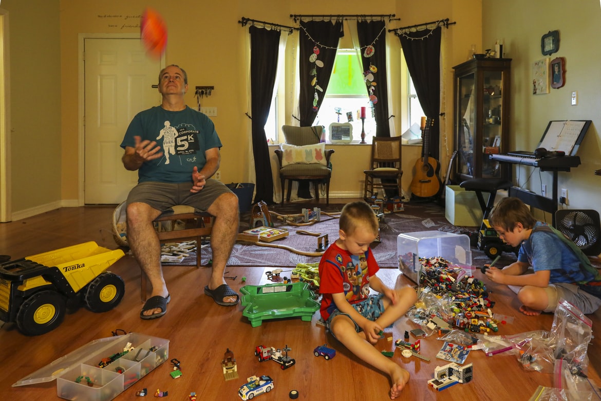 A father juggling while two boys play with toys spread across the living room floor